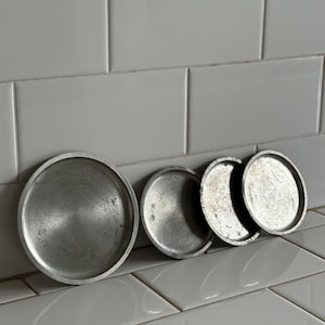 May include: A collection of five small, round, silver-toned metal plates. The largest plate has a textured, ridged edge. The plates are arranged on a white tiled surface, showcasing their simple, vintage design.