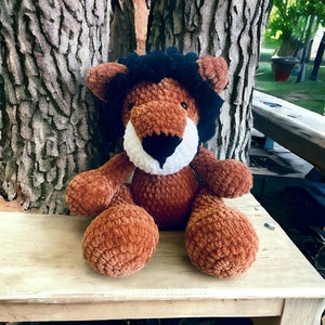 May include: A handmade plush lion toy with a brown body, black mane, and white and black facial features. The stuffed animal is sitting on a light-colored wooden surface. The lion has large, round paws and a friendly expression.