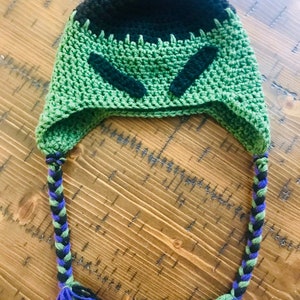May include: Green and black crocheted hat with long braided ties. The hat has a black face design with green eyes.