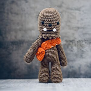 Op de afbeelding: Een bruin gebreid pluche speelgoed dat lijkt op een personage uit Star Wars met een oranje sjaal. Het speelgoed heeft een witte mond en zwarte ogen.