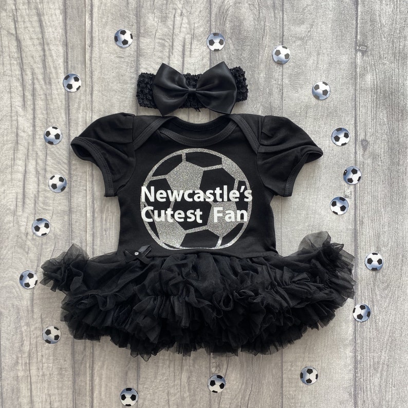 Black short sleeve tutu romper with silver football design and white text over the top saying Newcastle's Cutest Fan. Matching black bow headband above tutu romper.