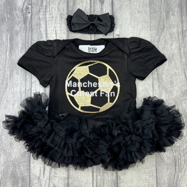 Baby girls black tutu romper with matching bow headband. Gold football design with white text saying Manchester's Cutest Fan. Manchester United, Man Utd, The Red Devils.