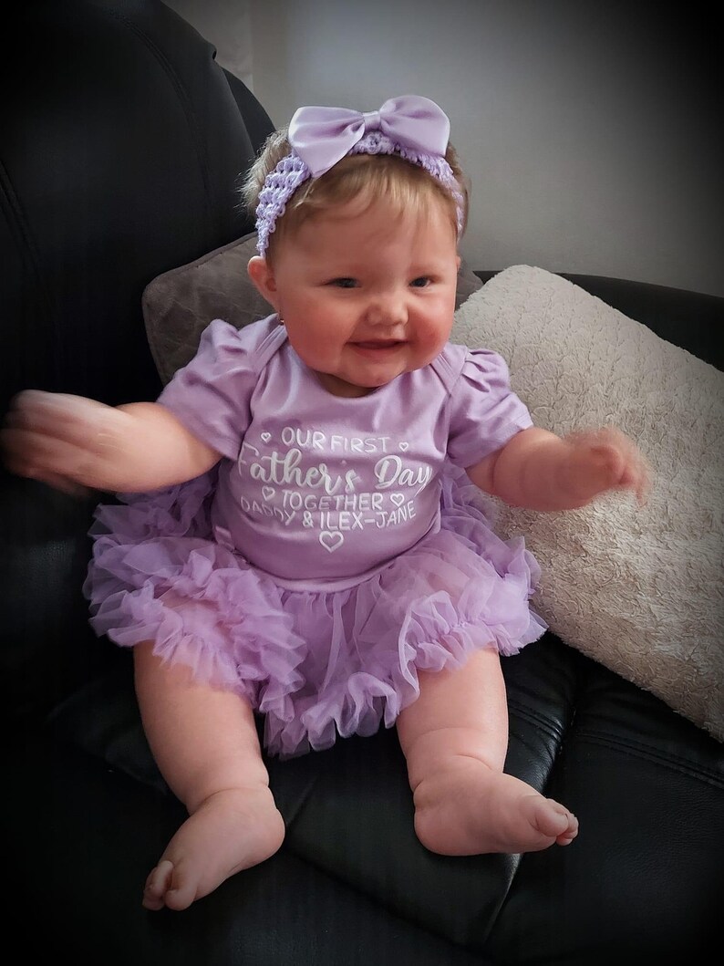 Baby Model wearing light purple short sleeve tutu romper with a matching bow and headband above. White love hearts design with lettering on the tutu romper saying Our First Father's Day Together Daddy & Daughter