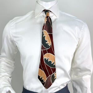 May include: A white long-sleeved dress shirt with a subtle textured pattern, paired with dark gray trousers and a necktie. The tie features a deep burgundy background with a repeating pattern of tan and dark green semi-circular shapes and thin white lines.
