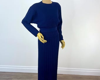 May include: A navy blue two-piece knit dress displayed on a mannequin. The long-sleeved top has a crew neck and a fitted waist. The skirt is pleated and reaches the ankles. The dress is made of a ribbed knit fabric.