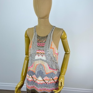 May include: A grey sequined vest top with a geometric pattern in pink, orange, and beige. The design features zigzags and curved lines. The top is displayed on a mannequin. The garment is embellished with sequins, creating a shimmering effect.