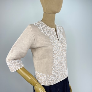 Op de afbeelding: Een beige cardigan met witte geborduurde details rond de halslijn, manchetten en zoom. De cardigan heeft een ritssluiting aan de voorkant.