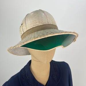 May include: A vintage safari hat with a wide brim and a green underside. The hat is made of woven straw with a brown band around the crown. The hat is displayed on a mannequin head, paired with a navy blue jacket.