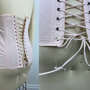 May include: A pale pink corset with a lace-up back and a floral pattern. The corset has a hook and eye closure at the front and is made of a soft, lightweight fabric.