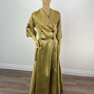 May include: An olive green vintage two-piece dress set displayed on a mannequin. The set comprises a double-breasted jacket with black buttons and a long skirt. The fabric has a subtle sheen, and the jacket features a wide collar and three-quarter sleeves.