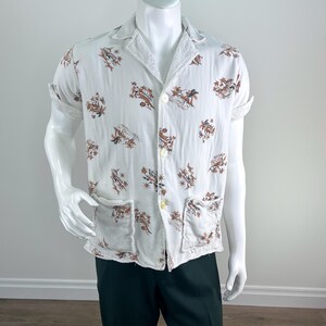 May include: White short-sleeved shirt with a brown and tan floral pattern. The shirt has a collar, two front pockets, and button closures. The sleeves are rolled up. The shirt is displayed on a mannequin.