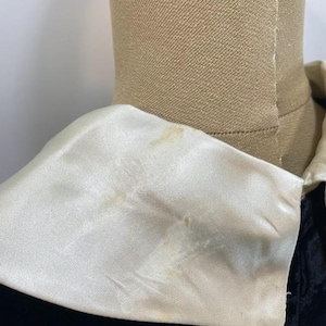 May include: A black garment featuring a large, wide, cream-colored satin collar. The collar has a smooth, reflective surface with visible light brown discoloration spots on the fabric. The garment is displayed on a tan-colored mannequin torso.