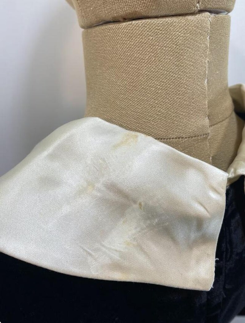 May include: A black garment featuring a large, wide, cream-colored satin collar. The collar has a smooth, reflective surface with visible light brown discoloration spots on the fabric. The garment is displayed on a tan-colored mannequin torso.