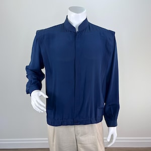 Puede incluir: Una chaqueta azul marino de manga larga con cuello mao, tapeta de botones oculta en la parte delantera y detalles de pliegues verticales a lo largo de los hombros. La chaqueta tiene un dobladillo ajustado y se muestra en un maniquí blanco.