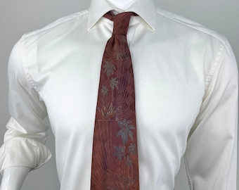 May include: A maroon necktie featuring a subtle checkered pattern and a printed design of palm trees and foliage in muted green and gold tones. The tie is knotted and worn over a crisp white long-sleeved dress shirt with a textured fabric finish.