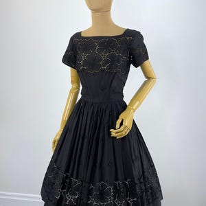 May include: A black dress featuring short sleeves, a square neckline, and a fitted bodice. The garment is adorned with intricate floral eyelet cutouts along the neckline and the hemline, complemented by a full, pleated skirt silhouette.