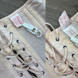 May include: A pink satin corset with white lace-up detailing. The corset has a label that reads "Charmode Made in U.S.A." and another label that reads "Style 257 S.F. Size 36 Rayon Rayon-E."