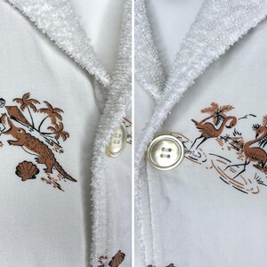 May include: White terry cloth robe with a tropical print featuring palm trees, a surfer, and an alligator. The robe has a shawl collar and large, round buttons.