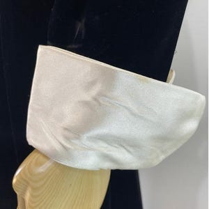 May include: A black velvet sleeve featuring a wide, contrasting white satin cuff. The cuff is smooth and has a slight sheen, creating a distinct visual separation from the dark, textured fabric of the sleeve. The garment is displayed on a mannequin.