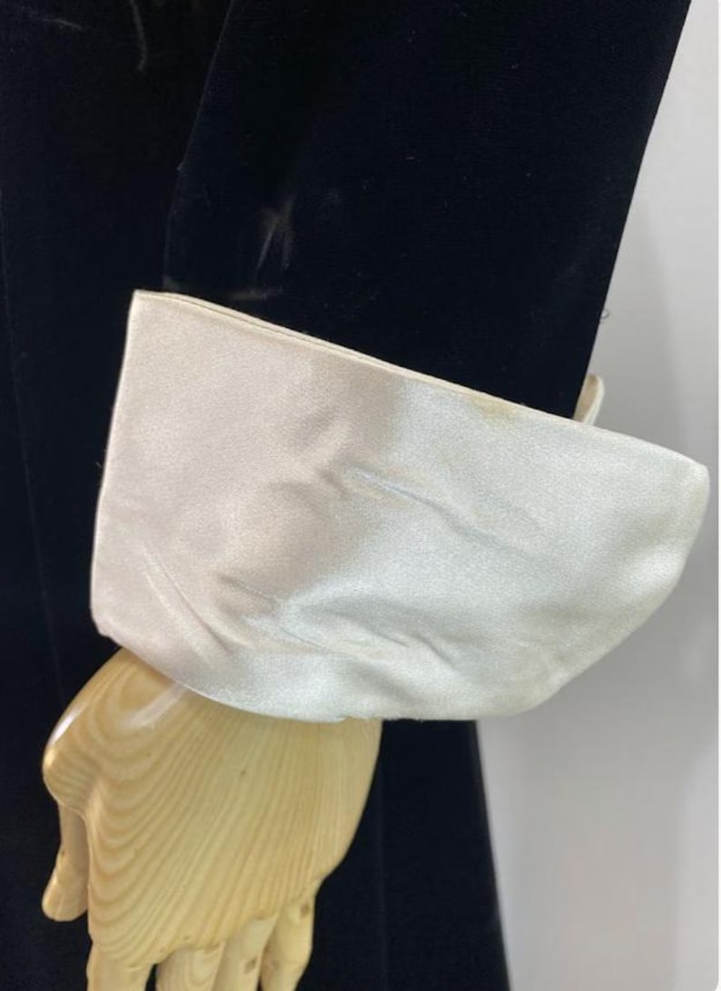 May include: A black velvet sleeve featuring a wide, contrasting white satin cuff. The cuff is smooth and has a slight sheen, creating a distinct visual separation from the dark, textured fabric of the sleeve. The garment is displayed on a mannequin.