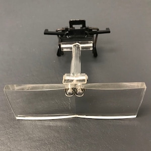May include: A pair of black and silver magnifying glasses with a clear plastic lens. The glasses are attached to a black metal frame with a clear plastic handle.