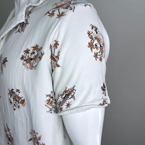 May include: White short-sleeved shirt with a brown and black tropical print featuring palm trees and figures. The shirt has a frayed collar and sleeve edges, giving it a casual look.