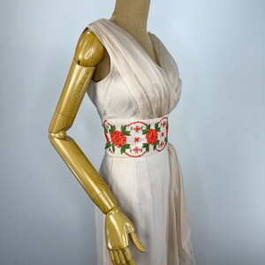 May include: A white sleeveless dress with a draped neckline and a floral embroidered belt. The dress has a fitted bodice and a flowing skirt.