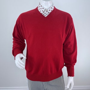 May include: A red V-neck sweater with long sleeves. Underneath, a white collared shirt with a burgundy polka dot pattern is visible. The mannequin is wearing striped pants.