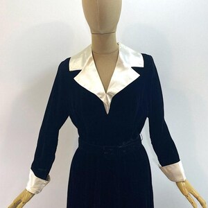 May include: A long-sleeved black velvet dress featuring a contrasting white satin notched collar and matching white satin cuffs. The garment includes a fitted waist with a matching black velvet belt, creating a structured silhouette on the mannequin.