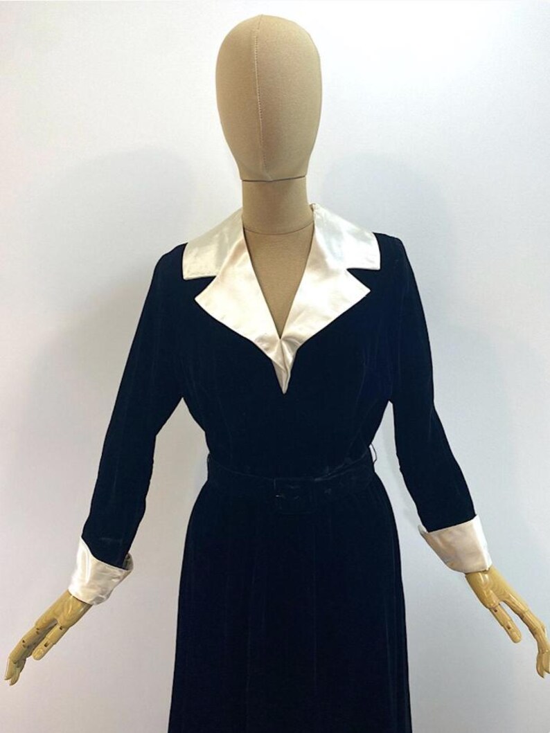 May include: A long-sleeved black velvet dress featuring a contrasting white satin notched collar and matching white satin cuffs. The garment includes a fitted waist with a matching black velvet belt, creating a structured silhouette on the mannequin.