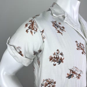May include: White short-sleeved shirt with a brown and black pattern featuring figures, palm trees, and other tropical elements. The shirt has a terry cloth collar and sleeve cuffs. The shirt is displayed on a mannequin.