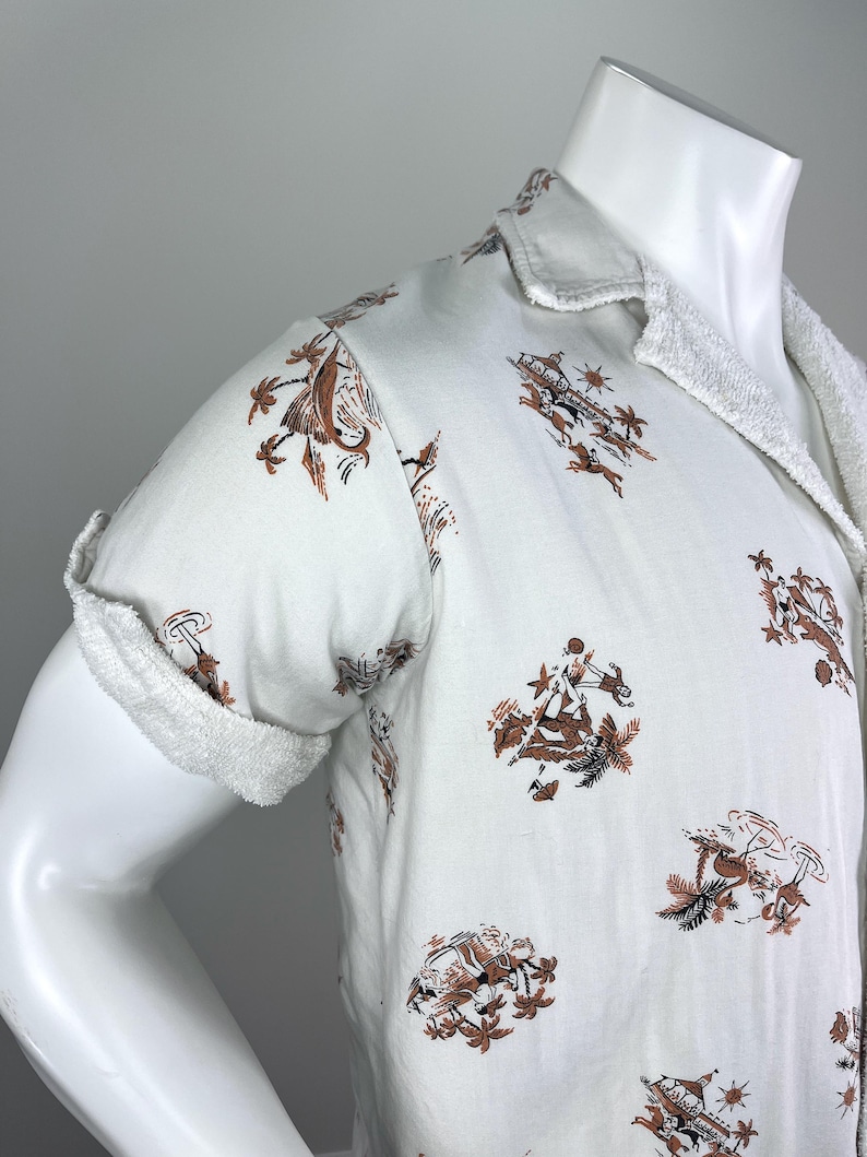 May include: White short-sleeved shirt with a brown and black pattern featuring figures, palm trees, and other tropical elements. The shirt has a terry cloth collar and sleeve cuffs. The shirt is displayed on a mannequin.