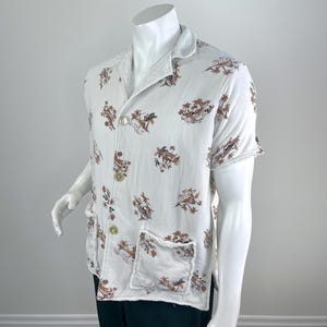 May include: White short-sleeved shirt with a brown and tan tropical print. The shirt has a collar, two front pockets, and two buttons. The edges of the shirt have a frayed design.