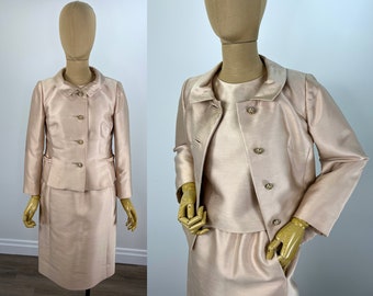 May include: A blush pink skirt suit with a matching jacket and skirt. The jacket has a collar and four buttons down the front. The skirt is knee-length and has a straight silhouette.