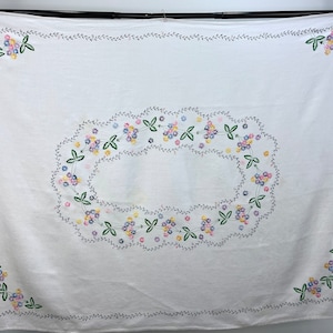 Puede incluir: Mantel de mesa de lino blanco con un delicado diseño de bordado floral. El bordado presenta una corona de pequeñas flores moradas, rosas y amarillas con hojas verdes, todo ello contorneado con hilo gris.