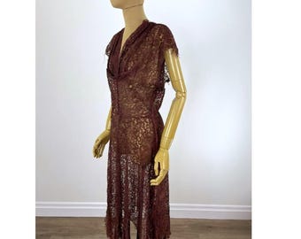 May include: A dark brown lace dress featuring a V-neckline, short sleeves, and a knee-length hem with a jagged, uneven edge. The garment is constructed from a sheer, intricate floral lace pattern that covers the entire surface of the dress.