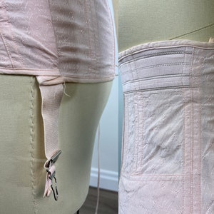 May include: A pink satin corset with a lace pattern and a garter strap on the side. The corset has a white elastic waistband.