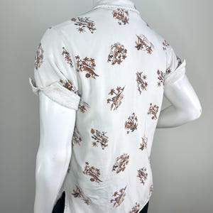May include: White short-sleeve shirt with a brown and tan tropical print. The shirt has a frayed collar and sleeve cuffs. The print features palm trees, people, and boats. The shirt is displayed on a mannequin.