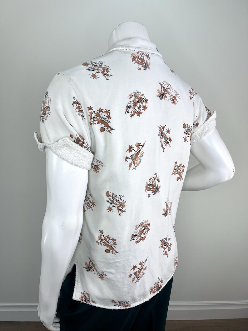 May include: White short-sleeve shirt with a brown and tan tropical print. The shirt has a frayed collar and sleeve cuffs. The print features palm trees, people, and boats. The shirt is displayed on a mannequin.