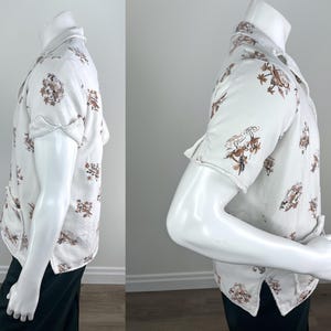May include: White button-down shirt with short sleeves, featuring a repeating brown floral and bird pattern. The shirt is displayed on a mannequin, showcasing its design and fit. The shirt has a classic collar and buttoned front.