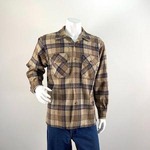 May include: A long-sleeve button-down shirt featuring a plaid pattern in shades of tan, brown, and dark blue. The shirt includes a standard collar, two chest pockets with buttoned flaps, and a full-length front button closure with dark buttons.