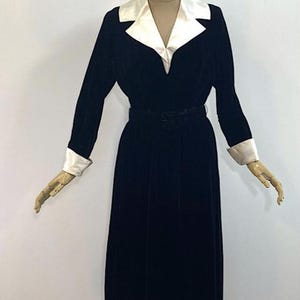 May include: A long-sleeved black velvet dress featuring a contrasting white satin notched collar and matching white satin cuffs. The garment includes a fitted waist with a matching black belt, a V-neckline, and a floor-length skirt silhouette.