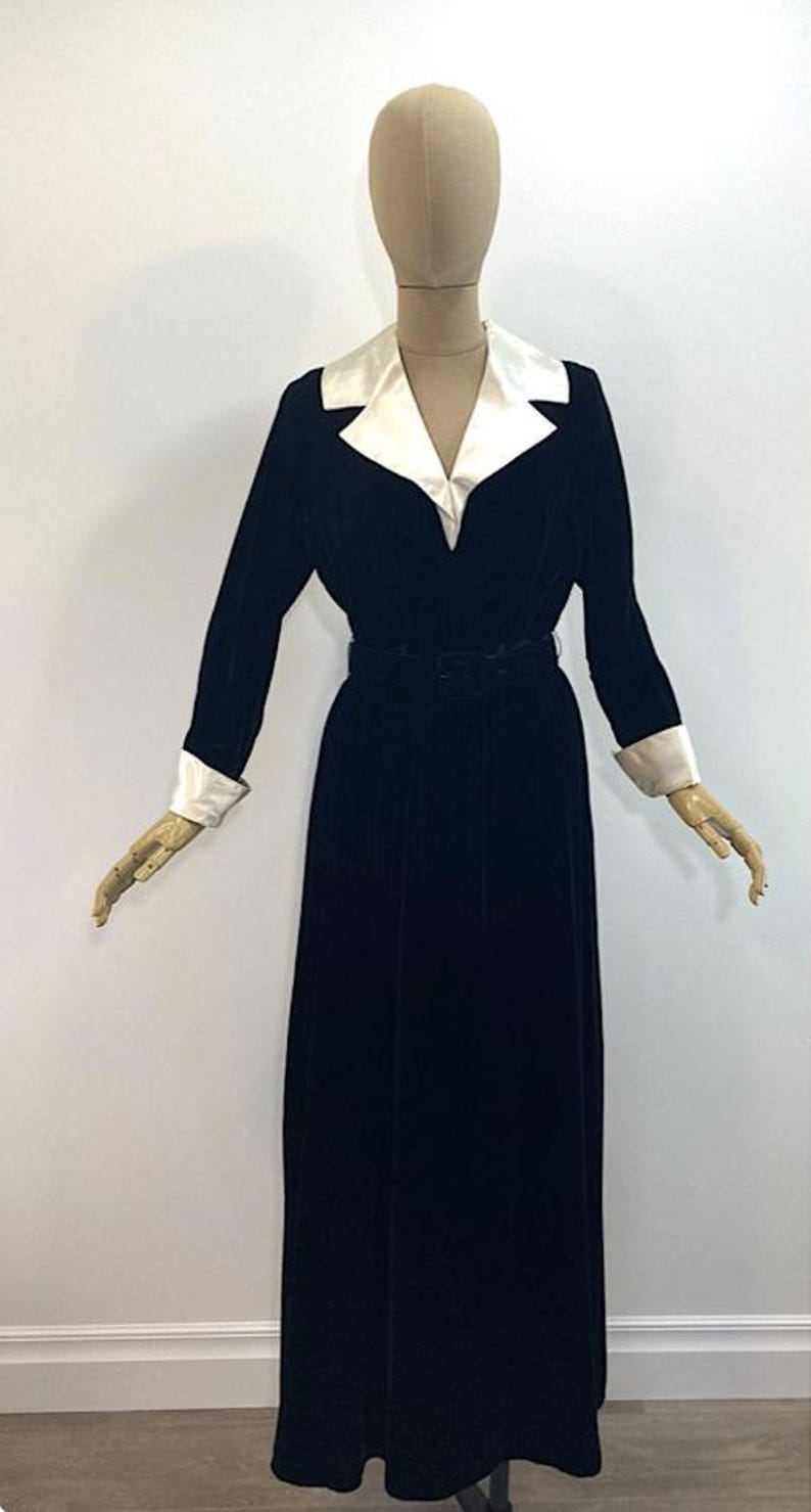 May include: A long-sleeved black velvet dress featuring a contrasting white satin notched collar and matching white satin cuffs. The garment includes a fitted waist with a matching black belt, a V-neckline, and a floor-length skirt silhouette.