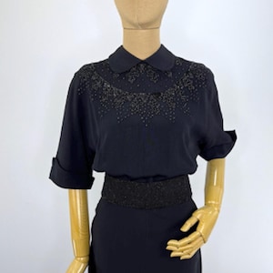 May include: A black blouse featuring a rounded collar and short sleeves with folded cuffs. The upper chest area is decorated with intricate black beadwork in a floral pattern. A wide, textured black waistband sits at the midsection.
