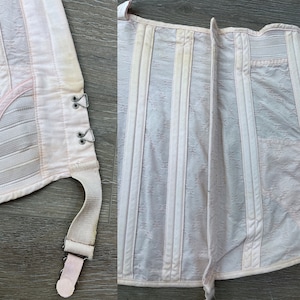 May include: A white corset with pink accents and a lace pattern. The corset has a hook and eye closure in the front and a lace-up back. The corset has a garter strap on the side.