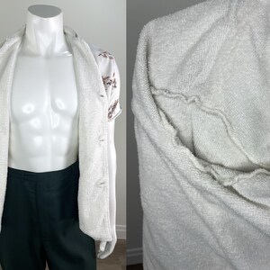 May include: A white terry cloth robe with a button-up shirt underneath. The shirt is white with a floral pattern and short sleeves. The robe is open, revealing the shirt and dark green pants.
