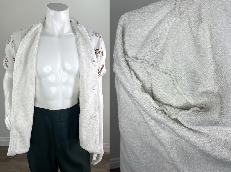 May include: A white terry cloth robe with a button-up shirt underneath. The shirt is white with a floral pattern and short sleeves. The robe is open, revealing the shirt and dark green pants.