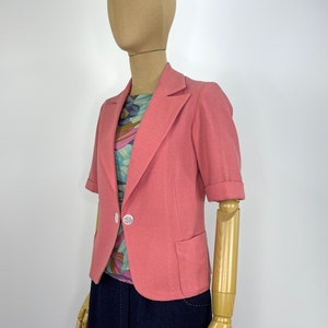 May include: A pink short-sleeved blazer with a single button closure and a pocket on the left side.