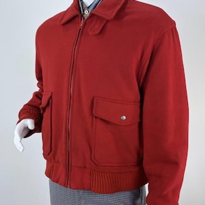 May include: A vibrant red bomber jacket featuring a classic fold-down collar, a full-length front zipper, and two large patch pockets with snap-button flaps. The garment includes ribbed knit cuffs and a ribbed hem, worn over a plaid shirt and trousers.