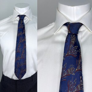 May include: A navy blue tie with a repeating pattern of brown and gold abstract shapes. The tie is displayed on a white dress shirt and is knotted at the collar. The shirt is on a mannequin.